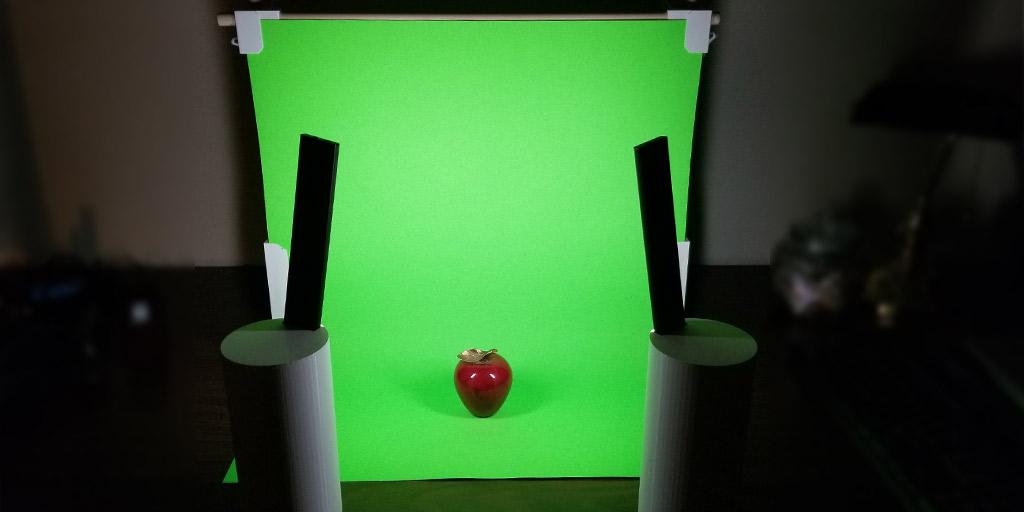 An image showing the entire 3D printed DIY photo booth backdrop setup. The DIY photo backdrop has the regular white poster board replaced with a green one to create a greenscreen effect and the LED light bar enclosure is being used to shine bright, soft light on the subject. The subject is the same apple as all previous photos show.